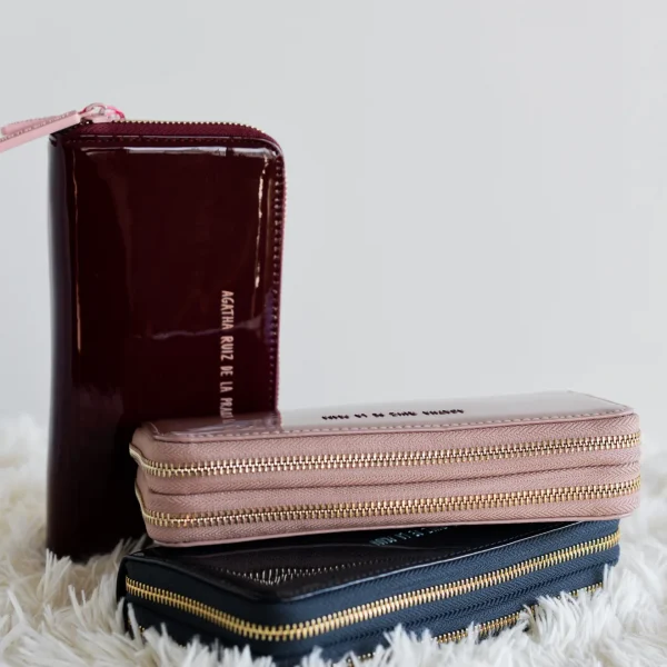 Espacio, orden y un acabado brillante que no pasa desapercibido. Este billetero grande de charol es perfecto para quienes necesitan capacidad sin renunciar al estilo. Cuenta con dos compartimentos independientes con cierre de cremallera, que facilitan la organización de tus tarjetas, billetes y monedas. El logo frontal en contraste aporta el toque distintivo de la colección, mientras que el diseño compacto asegura que puedas llevarlo cómodamente en cualquier bolso. Medidas: 19*10*5