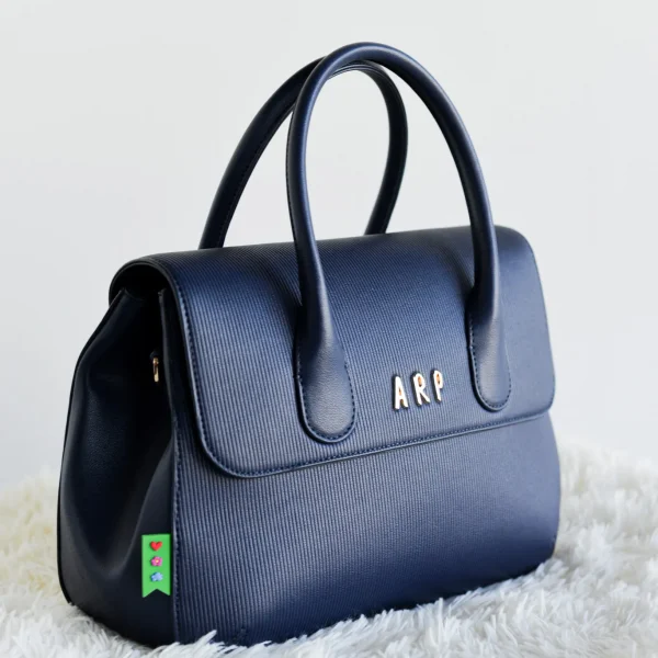 Elegancia y funcionalidad se unen en este bolso de mano con tapa, diseñado en piel vegana grabada con líneas verticales que aportan textura y un acabado sofisticado. Su formato compacto es ideal para quienes buscan un accesorio estiloso pero práctico. Incluye una correa desmontable, perfecta para llevarlo en la mano o colgado al hombro según la ocasión. Los detalles en dorado, como las iniciales ARP y el logo en la correa, aportan ese toque distintivo y exclusivo de Agatha Ruiz de la Prada. Medidas: 29*22*10