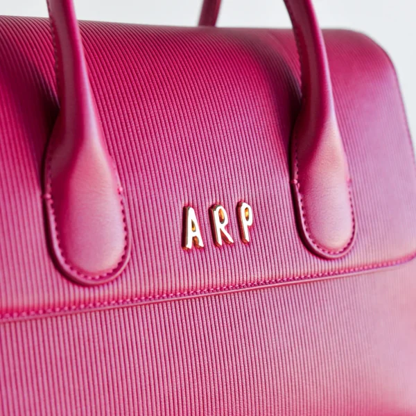 Elegancia y funcionalidad se unen en este bolso de mano con tapa, diseñado en piel vegana grabada con líneas verticales que aportan textura y un acabado sofisticado. Su formato compacto es ideal para quienes buscan un accesorio estiloso pero práctico. Incluye una correa desmontable, perfecta para llevarlo en la mano o colgado al hombro según la ocasión. Los detalles en dorado, como las iniciales ARP y el logo en la correa, aportan ese toque distintivo y exclusivo de Agatha Ruiz de la Prada. Medidas: 29*22*10