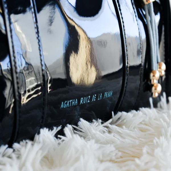 Este bolsito de mano cuadrado de charol es perfecto para quienes buscan un accesorio compacto pero con personalidad. El logo en contraste en el frontal y el pompón de cuerda a juego aportan el toque distintivo que marca la diferencia. Cuenta con cremallera superior para mayor seguridad, asas rígidas para llevarlo en la mano y una correa larga incluida que permite llevarlo al hombro o cruzado cuando quieras. Ideal para eventos, salidas o simplemente para sumar estilo a cualquier look. Medidas: 27*20*10