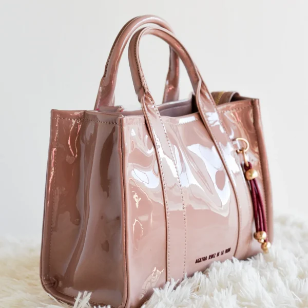 Este bolsito de mano cuadrado de charol es perfecto para quienes buscan un accesorio compacto pero con personalidad. El logo en contraste en el frontal y el pompón de cuerda a juego aportan el toque distintivo que marca la diferencia. Cuenta con cremallera superior para mayor seguridad, asas rígidas para llevarlo en la mano y una correa larga incluida que permite llevarlo al hombro o cruzado cuando quieras. Ideal para eventos, salidas o simplemente para sumar estilo a cualquier look. Medidas: 27*20*10