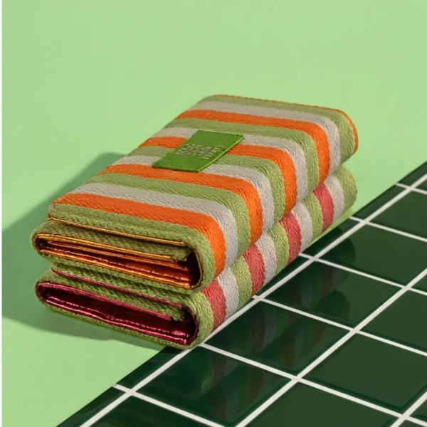 Este billetero con tapa pertenece a la colección en lona gruesa de líneas de color, un tejido resistente con el contraste vibrante que caracteriza a la línea: verde combinado con naranja o rosa fucsia. Su diseño es práctico, ligero y pensado para usar a diario sin renunciar al estilo. El billetero cuenta con cierre de clip en la tapa, que garantiza comodidad y seguridad al abrir y cerrar. En el interior incorpora un bolsillo con cremallera para monedas y varios departamentos independientes para organizar tarjetas, billetes y documentos. Todo pensado para que lleves lo necesario bien ordenado y accesible. Los detalles metalizados aportan un toque moderno que complementa perfectamente el look fresco y casual del tejido de lona. Medidas: 17*11*3